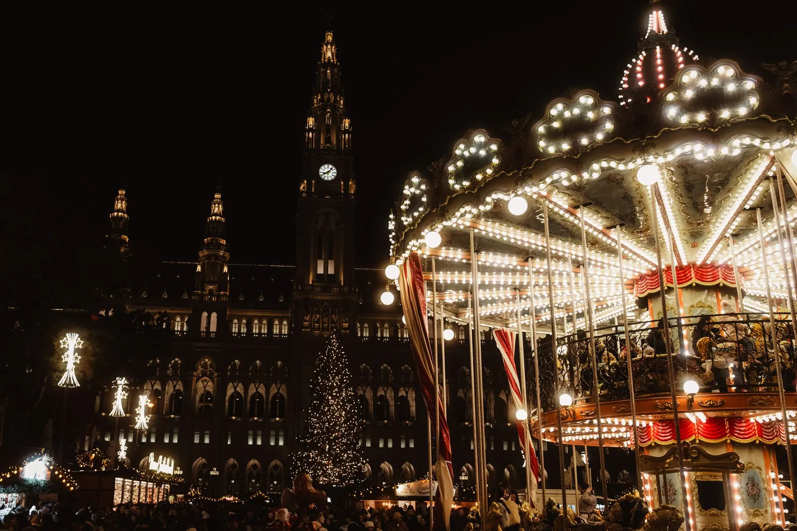 Brightly lit carousel at a Vienna Christmas market at night with City Hall towers behind