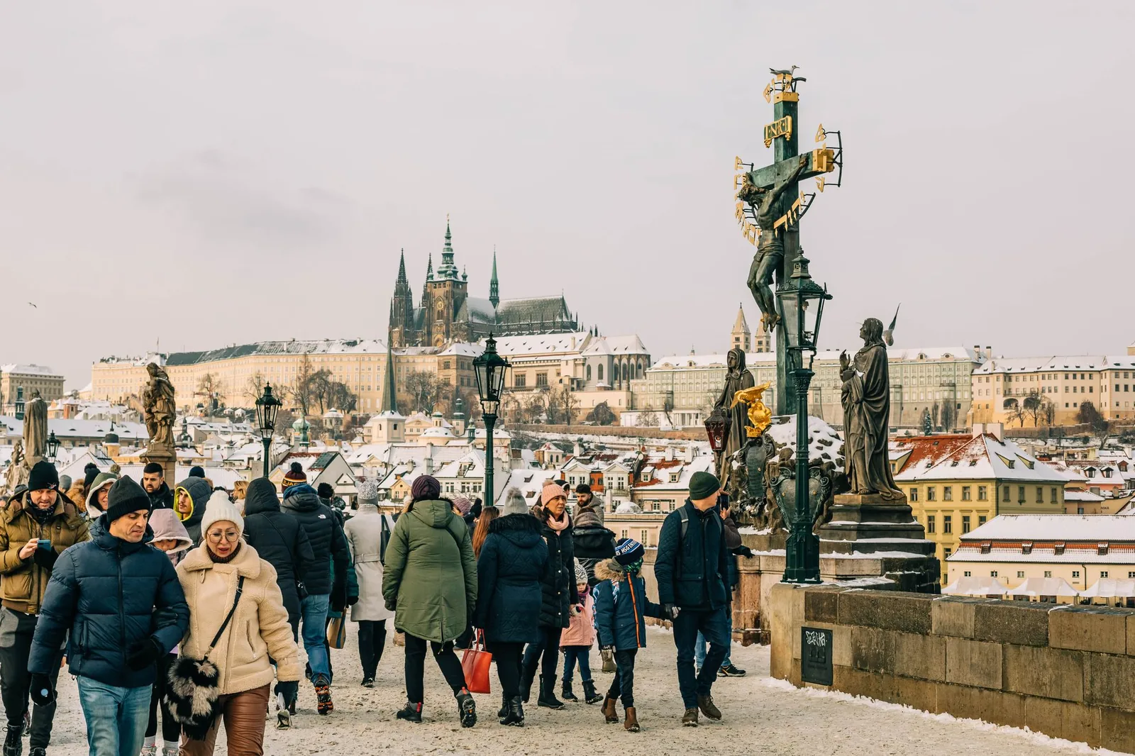 People walking across a snow-covered Charles Bridge with Prague Castle in the background